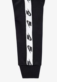 Fekete, karcsúsított szabású melegítőnadrág, fehér Nike márkajelzés szalaggal az oldalán és bordázott szegéllyel a bokáknál. Puha anyagtextúra.