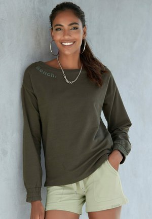 Olive green long-sleeve shirt with a relaxed fit and elastic cuffs, featuring a visible logo. Paired with light green shorts, casual design.