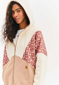Bluza z kapturem na zamek w kolorze kremowym, brązowym i różowym z cętkowanym wzorem. Posiada kaptur ze sznurkiem i przednie kieszenie. Miękka tekstura materiału.