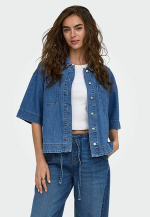 Jonge vrouw met lang golvend haar die een korte mouwen spijkerjasje draagt over een wit cropped topje en bijpassende spijkerbroek met een koord in de taille.