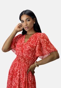 Red floral dress with short, flutter sleeves, gathered waist, and lightweight fabric. Features a v-neckline and a vibrant, eye-catching pattern.