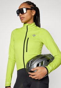 Kurtka rowerowa w neonowo zielonym materiale z wysokim kołnierzem i kieszenią na zamek, trzymana w dłoni szara kask. Smukły design, dopasowany krój.