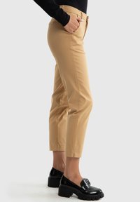 Kairi beiga kokvilnas maisījuma bikses līdz potītēm ar šauru piegriezumu, sānu kabatām un gludu faktūru, apvienotas ar melniem masīviem mokasīniem.