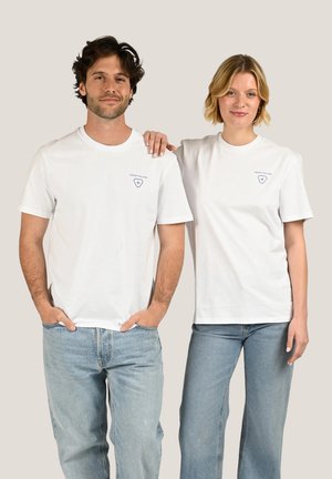 Homme et femme debout, tous deux portent des t-shirts blancs avec un logo bleu et des jeans bleu clair, la femme posant la main sur l'épaule de l'homme.