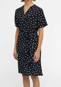 Robe marine avec des pois blancs, à manches courtes, décolleté en V et ceinture nouée à la taille ; fabriquée en tissu lisse avec une coupe décontractée.
