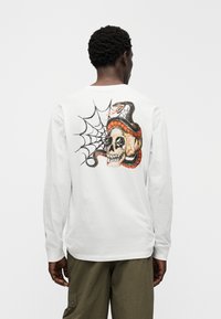 Λευκό μακρυμάνικο μπλουζάκι με πολύχρωμο γραφικό στο πίσω μέρος που απεικονίζει κρανίο, φίδι και σχέδιο ιστού αράχνης. Μαλακό βαμβακερό ύφασμα, κανονική γραμμή.