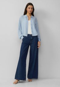 Blauw-wit gestreepte button-up shirt gecombineerd met donkere wijde spijkerbroeken en zilveren hakken. Het shirt heeft een ontspannen pasvorm en manchetten.