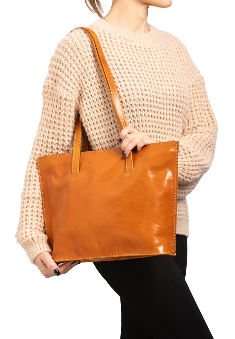 Borsa tote in pelle marrone chiaro con una finitura liscia e lucida, manici corti e dettagli di cucitura visibili. Indossata da una persona su uno sfondo neutro.