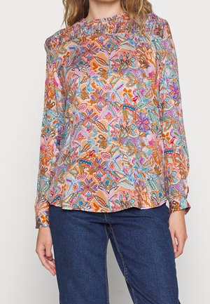 Donna che indossa una blusa a maniche lunghe con un motivo floreale astratto e colorato e jeans di denim blu, in piedi su uno sfondo chiaro.