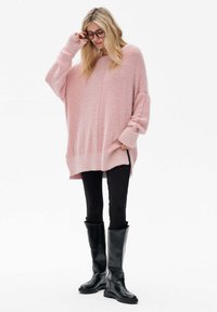 Chunky, oversized roze gebreide trui met ribbelzoom en -manchetten, gecombineerd met zwarte leggings en zwarte leren laarzen tot de knie.