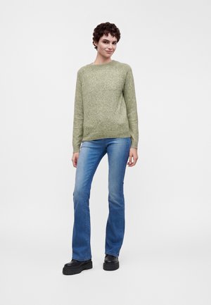 Person stående iført en grøn strikket sweater, blå vide jeans og sorte platformsko mod en ensfarvet hvid baggrund.