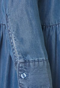 Chemise en denim à manches légères de couleur bleu clair, dotée d'un design élastique froncé, d'une fermeture à boutons et d'une couture détaillée le long de la couture.