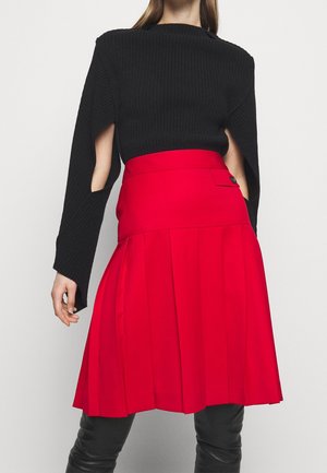 Haut noir à rayures avec des manches longues et des découpes, associé à une jupe plissée rouge avec une poche latérale et des détails de boutons.