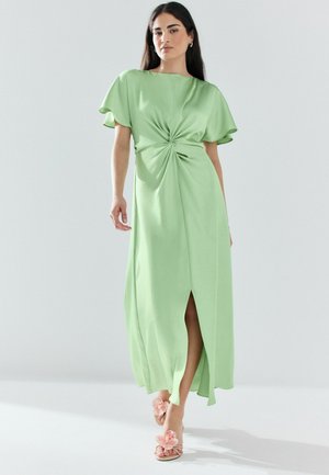 Hosszú sötét hajú nő világoszöld midi ruhában, melynek elöl csavart részlete és fodros ujjai vannak, rózsaszín virágmintás, magassarkú szandállal párosítva.