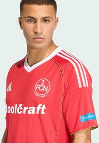 Camiseta de fútbol roja con acentos blancos, diseño de cuello en V, que presenta el logo y el texto "1. FCN". Hombros a rayas, ribete dorado y logotipos de patrocinadores.