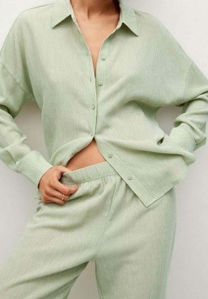 Personne portant une chemise boutonnée texturée vert clair et un pantalon assorti avec taille élastique, ajustant la taille du pantalon d’une main.