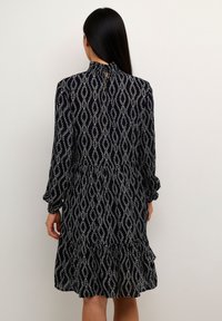 Zwarte jurk met wit golfpatroon, lange mouwen, geplooide zoom en een keyhole-detail aan de achterkant. Gemaakt van lichtgewicht stof, tot de knie rekkend.