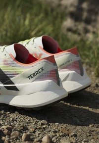 Paar weiße und orange Terrex-Laufschuhe, die auf felsigem Boden mit grünem Gras im Hintergrund liegen.