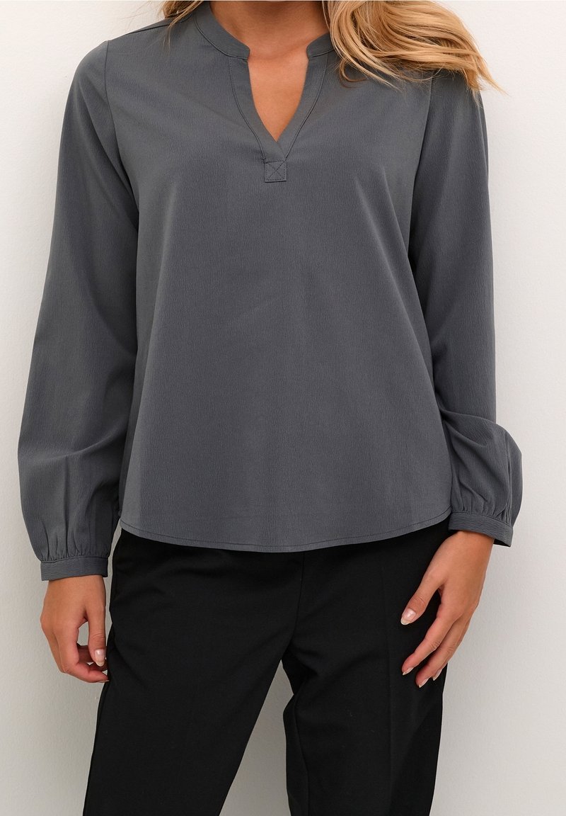 Blouse grise à manches longues avec un col en V peu profond, tissu texturé et poignets froncés. Assortie à un pantalon noir, design minimaliste et ajusté.