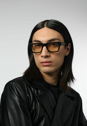 Des lunettes noires en forme de rectangle avec des verres ambrés, portées par une personne ayant de longs cheveux noirs, vêtue d'une veste en cuir noire, sur un fond gris.