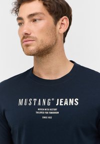 Tmavě modré bavlněné tričko s bílým grafickým textem "MUSTANG JEANS", kulatým výstřihem a krátkými rukávy.