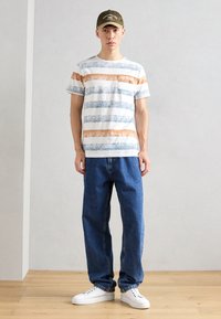 Camiseta blanca con rayas horizontales azules y naranjas, con detalle de bolsillo. Combinada con unos vaqueros azules de corte holgado y zapatillas blancas. Gorra oliva.