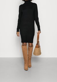 Svart polotröja klänning med långa ärmar, knälång siluett. Kombinerad med tanfärgade knähöga stövlar och en vävd tote-väska. Minimalistisk design.