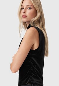 Blonde vrouw met steil haar die een mouwloos zwart fluwelen top draagt, zijwaarts staat en naar de camera kijkt tegen een effen achtergrond.
