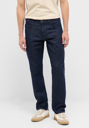 Mann trägt dunkelblaue Straight-Leg-Jeans und beige Sneakers, steht vor einem schlichten hellen Hintergrund.