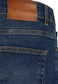 Tamno plave traperice imaju pravokutnu smeđu kožnu oznaku s natpisom "2Y PREMIUM" na struku, s vidljivim šavovima i džepovima.