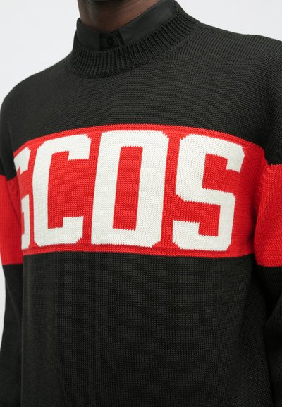 Suéter preto de malha com uma faixa horizontal vermelha e grandes letras brancas "GCDS" no peito, usado sobre uma camisa preta de colarinho.