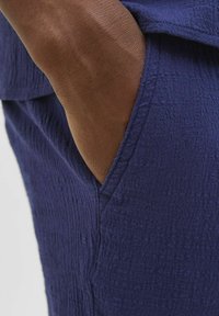 Tissu texturé bleu marine avec un motif de fronce subtil, comportant une poche latérale et une manche lisse en contraste avec le vêtement.