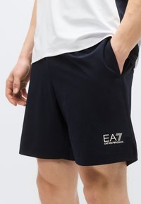 Tmavomodré športové šortky z ľahkého materiálu, s bočnými vreckami a bielym logom EA7 na ľavom stehne. Hladká textúra.