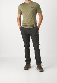 Zelené tričko s krátkým rukávem a kulatým výstřihem, černé logo; tmavé, úzké kalhoty; odolné boty s hnědými podrážkami.