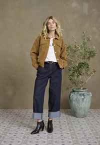 Tan suède jas, witte shirt, donkere denim broek met omgeslagen broekspijpen, zwarte haklaarzen, staande naast een decoratieve groene vaas met een olijftak.