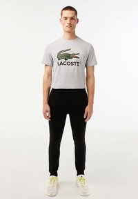 Grijs katoenen T-shirt met een groen krokodillenlogo en de tekst "LACOSTE"; gecombineerd met zwarte strakke broek en witte sneakers met gele accenten.
