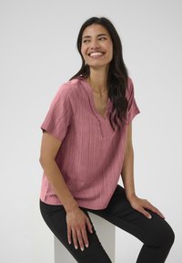Blouse rose à manches courtes avec une finition texturée, dotée d'un col en V et de détails festonnés au niveau du cou, associée à un pantalon noir.