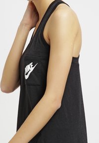 Černý top bez rukávů vyrobený z měkké tkaniny s kulatým výstřihem a kapsou s logem na levé straně, obsahující bílé logo Nike.