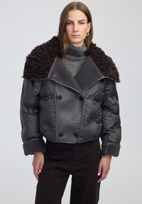 Kobieta w szarej pikowanej kurtce z dużym teksturowanym kołnierzem, szarym swetrze z golfem i ciemnych spodniach, stojąca na jednolitym tle.