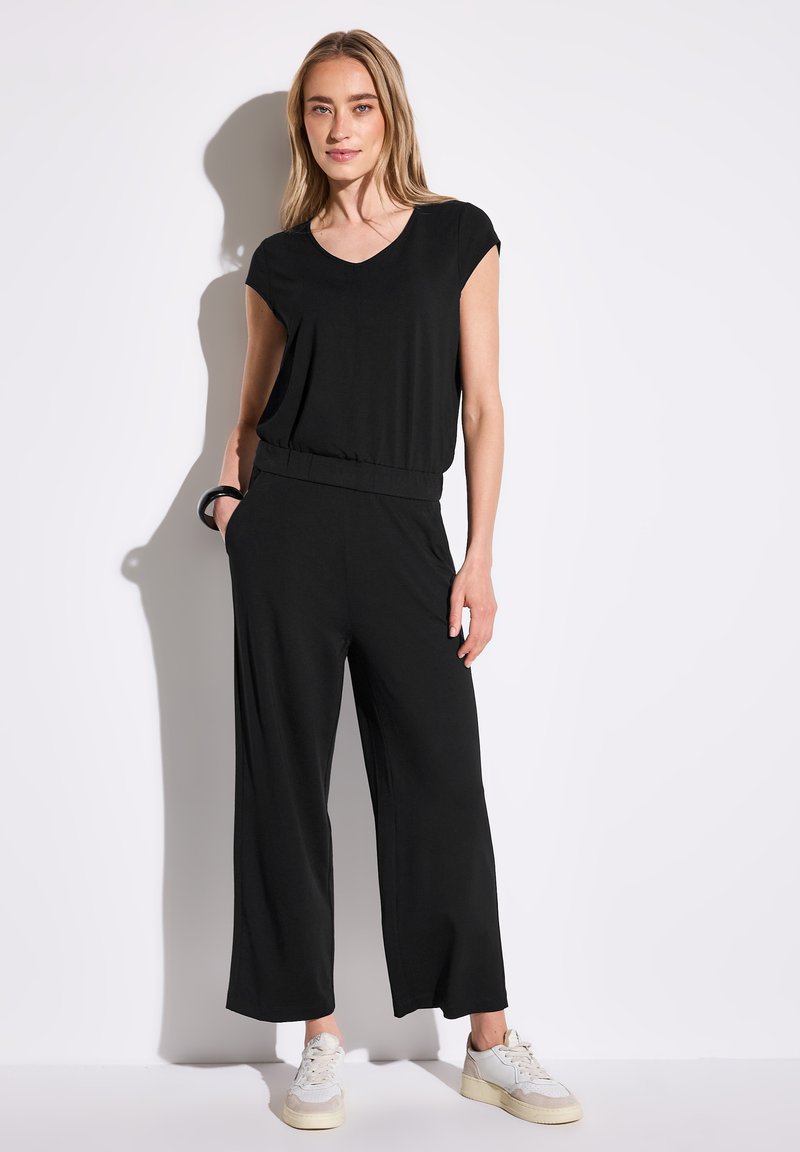 Mono negro con mangas cortas, escote en V, cinturilla elástica y diseño de pierna ancha. Combinado con zapatillas deportivas blancas.