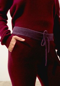 Bordowy sweter dzianinowy o prążkowanej teksturze, z fioletowym sznurkiem w talii i kieszenią po lewej stronie koordynujących spodni.