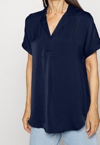 Blusa a maniche corte blu navy con scollatura a V e texture sottile; vestibilità ampia e orlo arrotondato. Abbinata a jeans in denim blu chiaro.