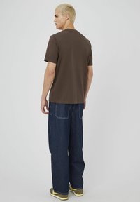 T-shirt marron à manches courtes avec un col rond, coupe décontractée et fentes latérales, associé à un jean en denim bleu ample et des baskets vertes et jaunes.
