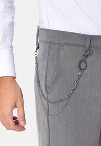 Pantaloni grigi con una finitura testurizzata presentano un accessorio a catena metallica attaccato a un passante della cintura, completato da un dito vicino alla tasca.