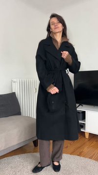 Fekete hosszú kabát övvel, dupla soros gombokkal és széles gallérral, barna kötött pulóver és barna kordbársony nadrág fölött. Fekete cipő.