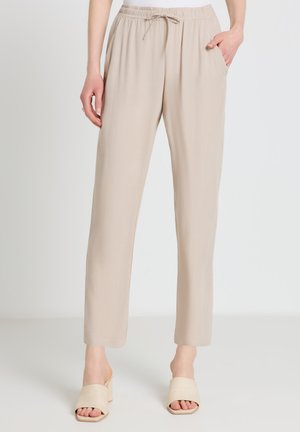 Beige high-waist drawstring pants with side pockets worn by a person, paired with light beige heeled mules, standing on a white floor.