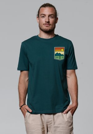Teal short-sleeve T-shirt in soft fabric, featuring a colorful mountain and sun graphic on the left chest area, with "GOOD VIBE" text.