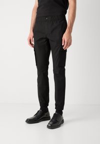 Pantaloni cargo negri cu talie elastică, două buzunare laterale și manșete la glezne. Purtați cu pantofi negri cu șireturi.
