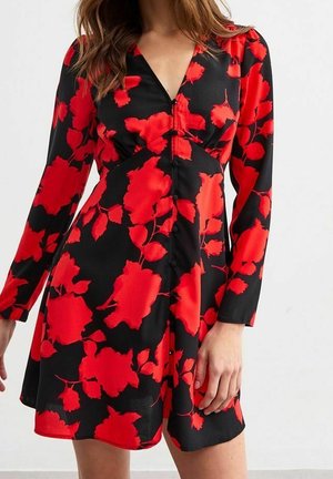 Kvinde iført en sort kjole med store røde blomsterprint, lange ærmer og V-udskæring, stående mod en ensfarvet lys baggrund.