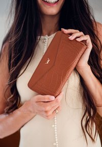 Brown, crocodile-embossed wallet with gold hardware detail, rectangular shape, and smooth texture, held in a hand against a light background.